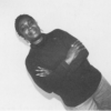 A black and white self-portrait of Jacqueline, a Black women artist, shot at an angle with her hands crossed between her arms. She is wearing a black turtleneck, jeans, and drooping earrings, and has a bright, slightly-opened smile with kind eyes.