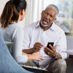 Frustrated senior man holds smartphone while discussing an issue with a female therapist.