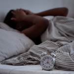 Sleepless, woman suffering from insomnia, sleep apnea or stress, headache or migraine. Tired and exhausted african american lady awake in middle of night, near alarm clock with time in dark, panorama