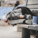 Poor homeless man or refugee sleeping on the wooden bench on the urban street in the city,